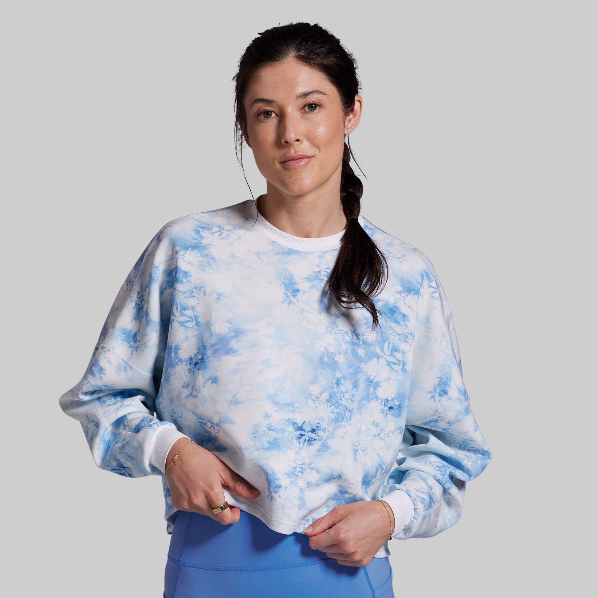 A woman modeling a light blue and white tie-dye crewneck sweatshirt, paired with blue pants, against a solid light gray background.