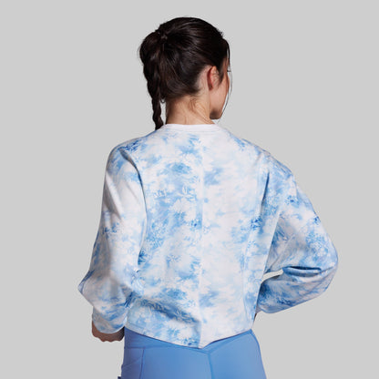 A woman shown from behind wearing a white long-sleeve sweatshirt with a light blue tie-dye pattern, paired with light blue pants, against a solid gray background.