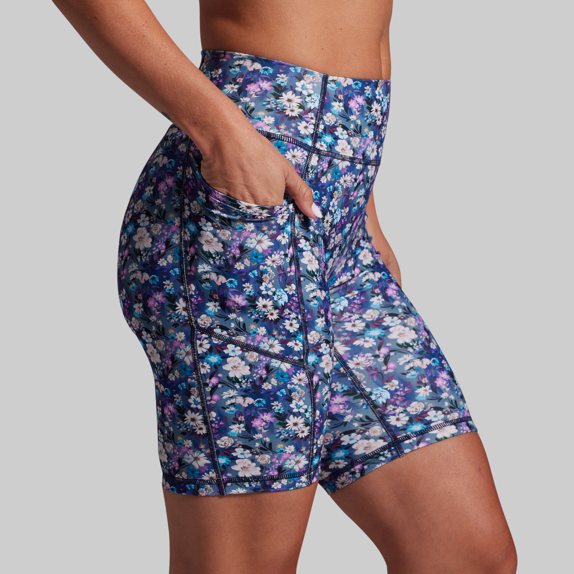 A close-up of a model wearing high-waisted athletic biker shorts with a blue, purple, and white floral pattern, featuring side pockets and dark contrast stitching, set against a solid gray background.