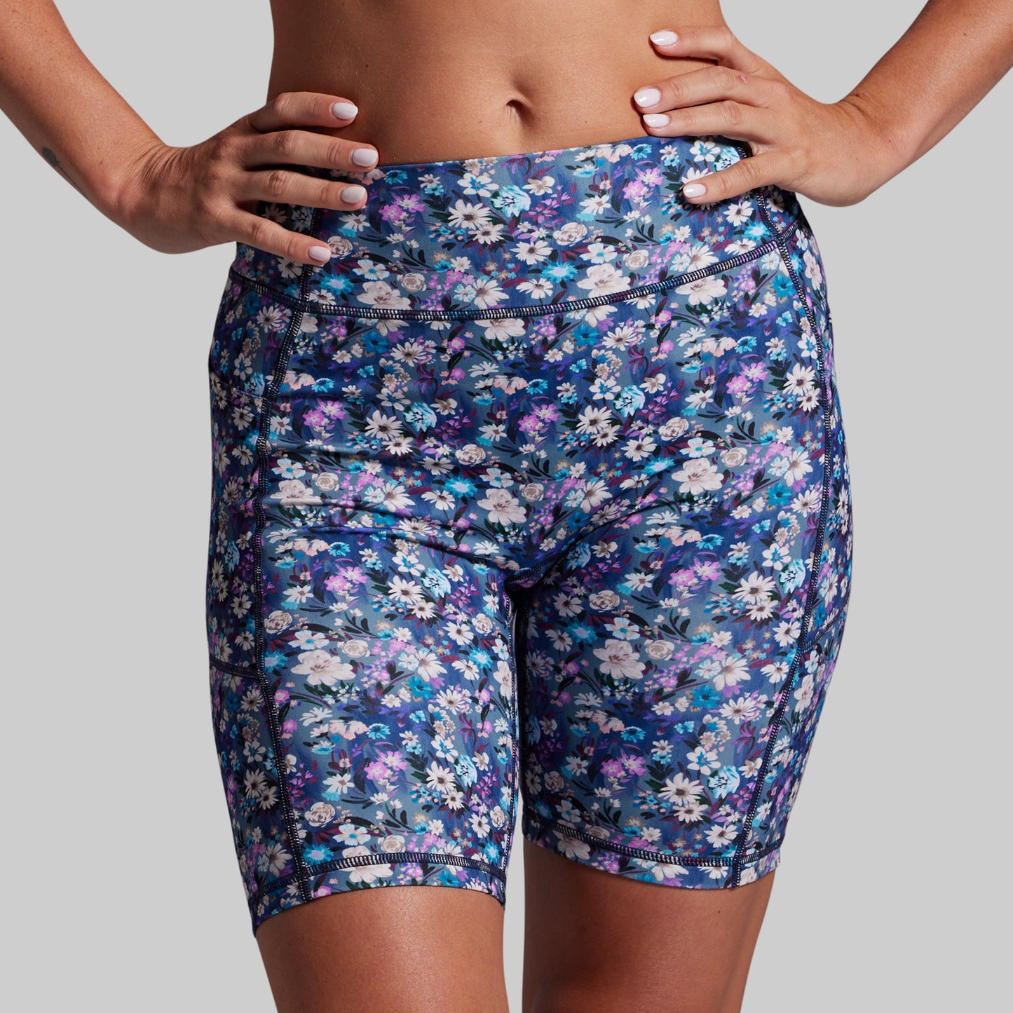 A close-up of a model wearing high-waisted athletic biker shorts with a dense floral print in navy, blue, purple, and white, set against a solid gray background.