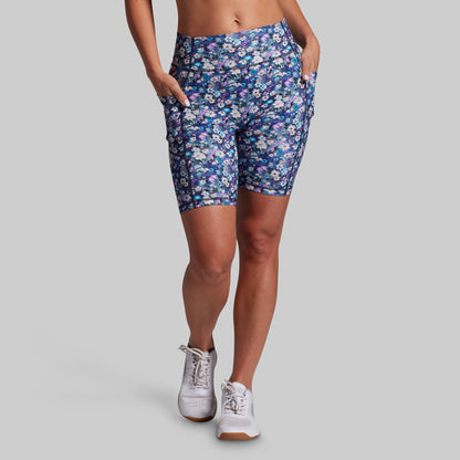 A model wearing blue floral patterned bike shorts with side pockets and white sneakers, shown from the waist down against a solid gray background.