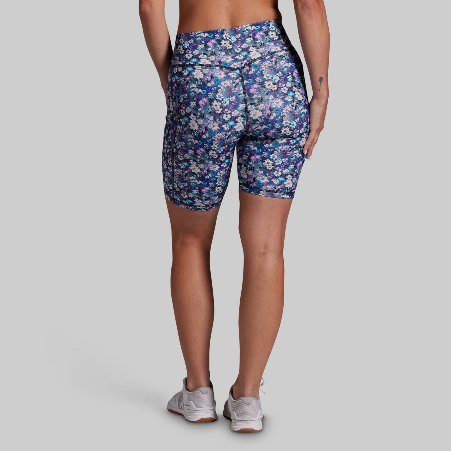Rear view of a model wearing floral print biker shorts in blue, purple, and white, paired with light gray sneakers, against a solid gray background.