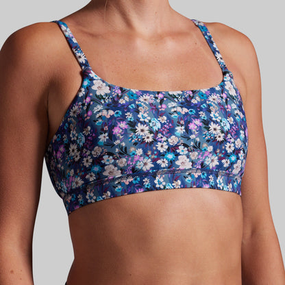 A close-up of a woman wearing a floral sports bra with blue, purple, and white flowers on a dark background, set against a solid gray studio backdrop.