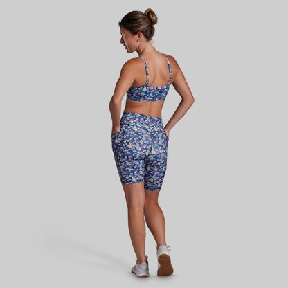 A woman is shown from the back wearing a matching athletic set consisting of a blue floral sports bra and biker shorts, set against a solid gray background.