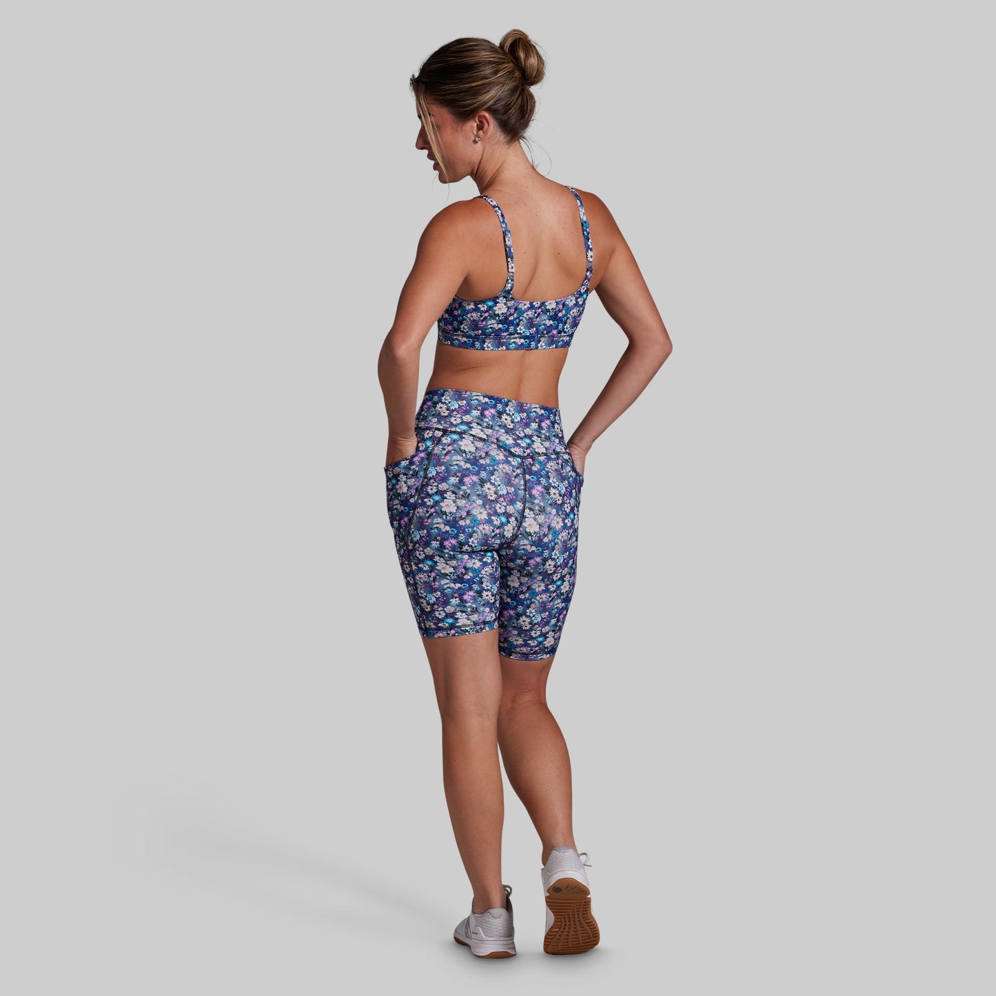 A woman is shown from the back wearing a matching athletic set consisting of a blue floral sports bra and biker shorts, set against a solid gray background.