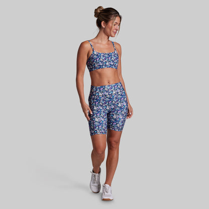 A woman modeling a matching athletic set consisting of a blue floral print sports bra and high-waisted biker shorts, paired with white sneakers, against a solid gray background.