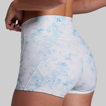 Close-up back view of a model wearing high-waisted athletic shorts with a light blue and white tie-dye pattern, set against a solid gray background.