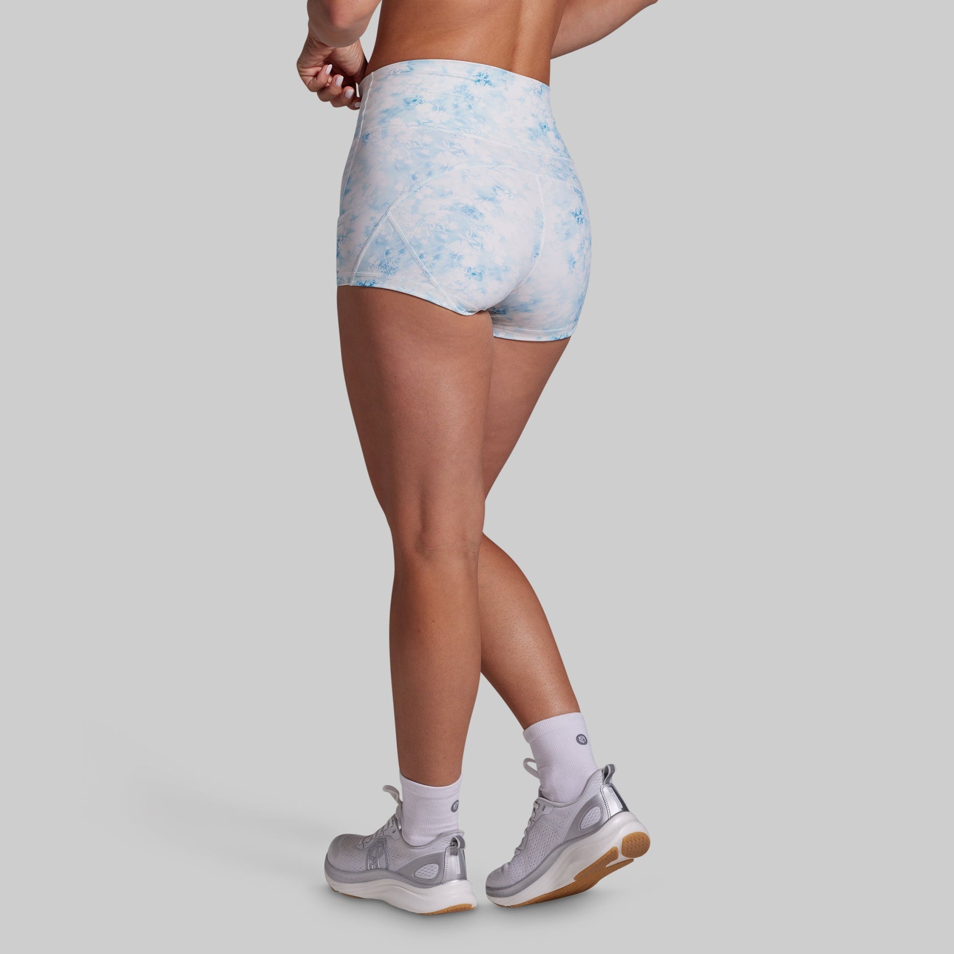 Rear three-quarter view of a model wearing high-waisted athletic shorts with a light blue and white watercolor pattern, paired with white socks and gray running shoes, against a solid gray background.