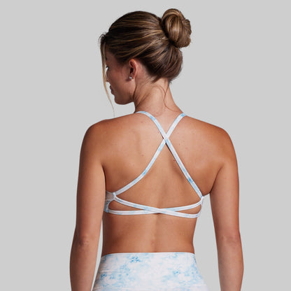 A woman shown from the back wearing a light blue and white tie-dye sports bra with crisscrossing straps, paired with matching tie-dye bottoms, against a solid gray background.