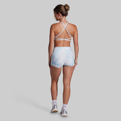 A woman shown from the back wearing a white sports bra with crisscross straps and light blue and white patterned athletic shorts, standing against a solid gray background.