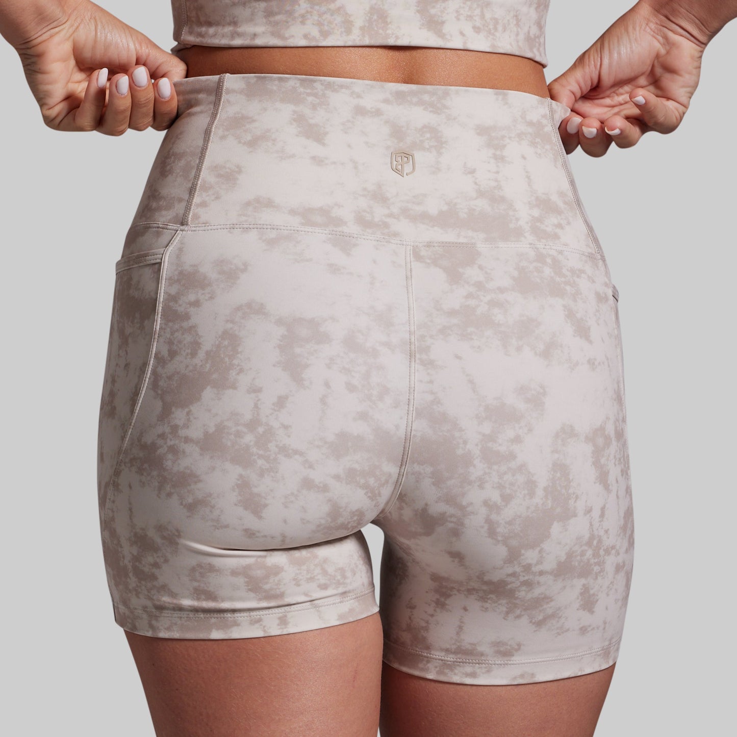 Rear view of a person wearing high-waisted athletic shorts with a light beige and taupe mottled pattern, holding the waistband with both hands against a light gray background.