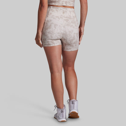 Rear view of a model wearing high-waisted athletic shorts with a light beige and gray abstract tie-dye pattern, paired with white athletic shoes, against a solid gray studio background.