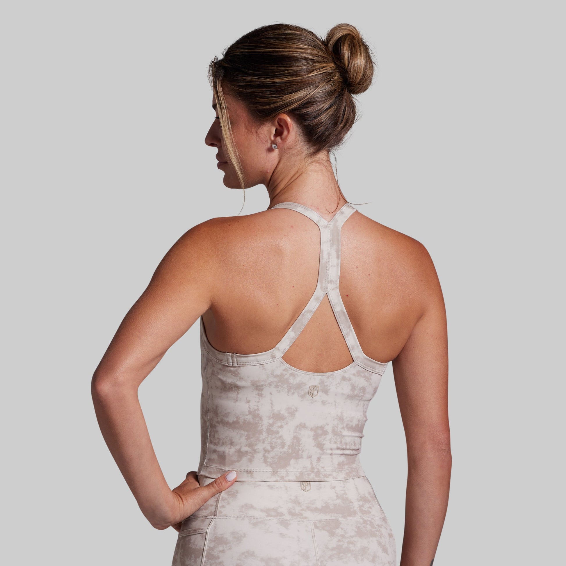 A woman shown from the back wearing an extended sports bra with racerback straps and a matching high-waisted shorts, both in a light beige and white tie-dye pattern, against a solid gray background.