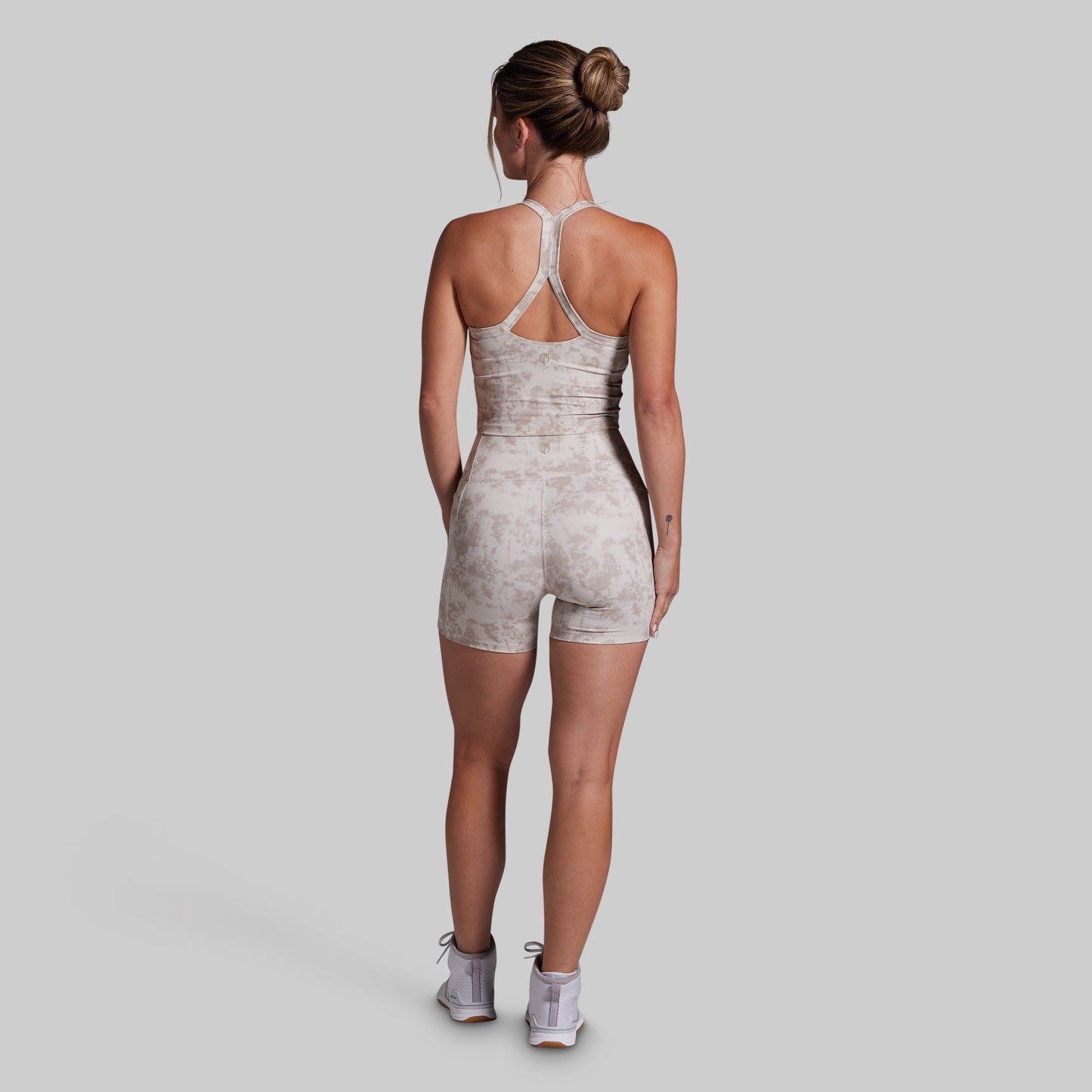 A woman viewed from behind wearing a light beige and white marbled athletic sports bra and shorts set with a crossover back strap, paired with white sneakers, against a solid gray background.
