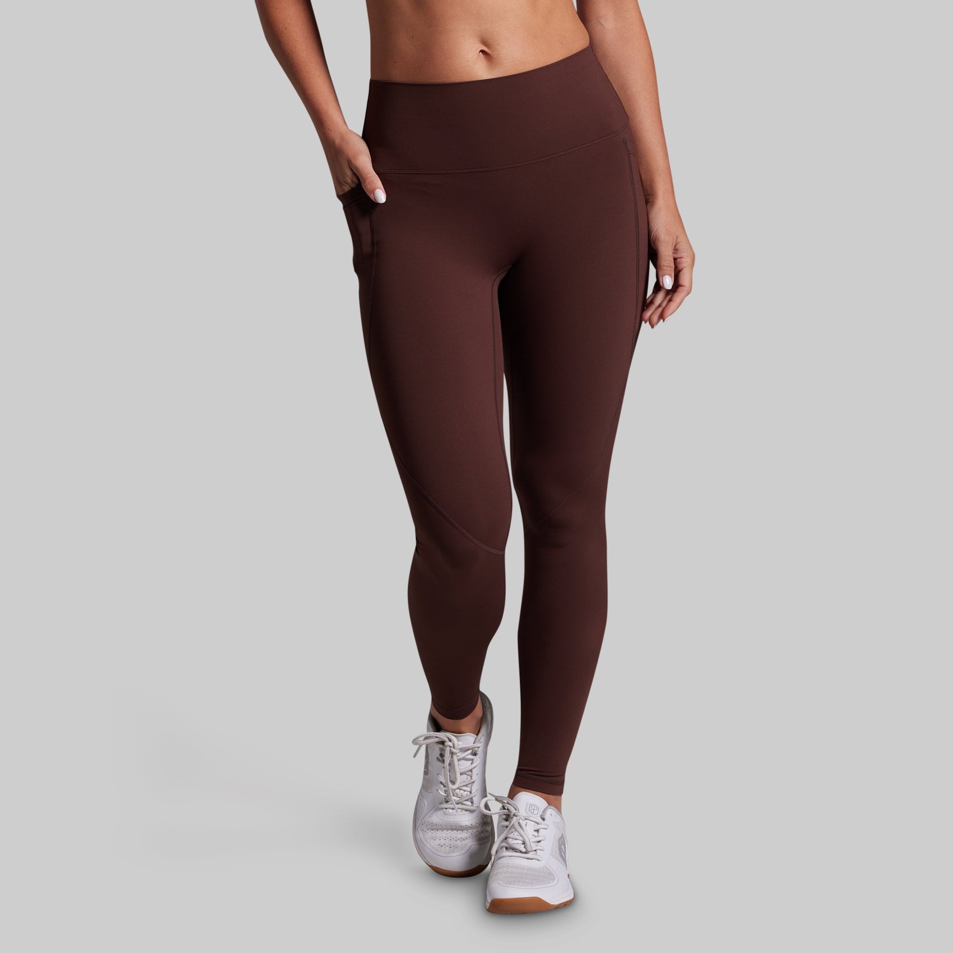 A model wearing high-waisted, dark brown athletic leggings with side pockets, paired with white sneakers, shown from the waist down against a solid gray background.