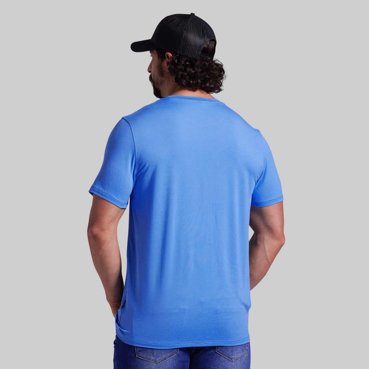 A man viewed from behind wearing a bright blue short-sleeve crewneck t-shirt, a black baseball cap, and blue jeans, set against a solid light gray background.
