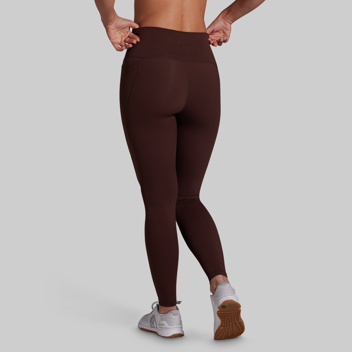 A person shown from the back wearing dark brown high-waisted leggings and white athletic sneakers against a light gray studio background.