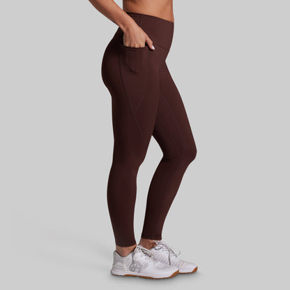 A person wearing high-waisted, form-fitting leggings in a dark brown color, shown from the waist down against a solid light gray background. The leggings feature side pockets, and the person is wearing white athletic sneakers.