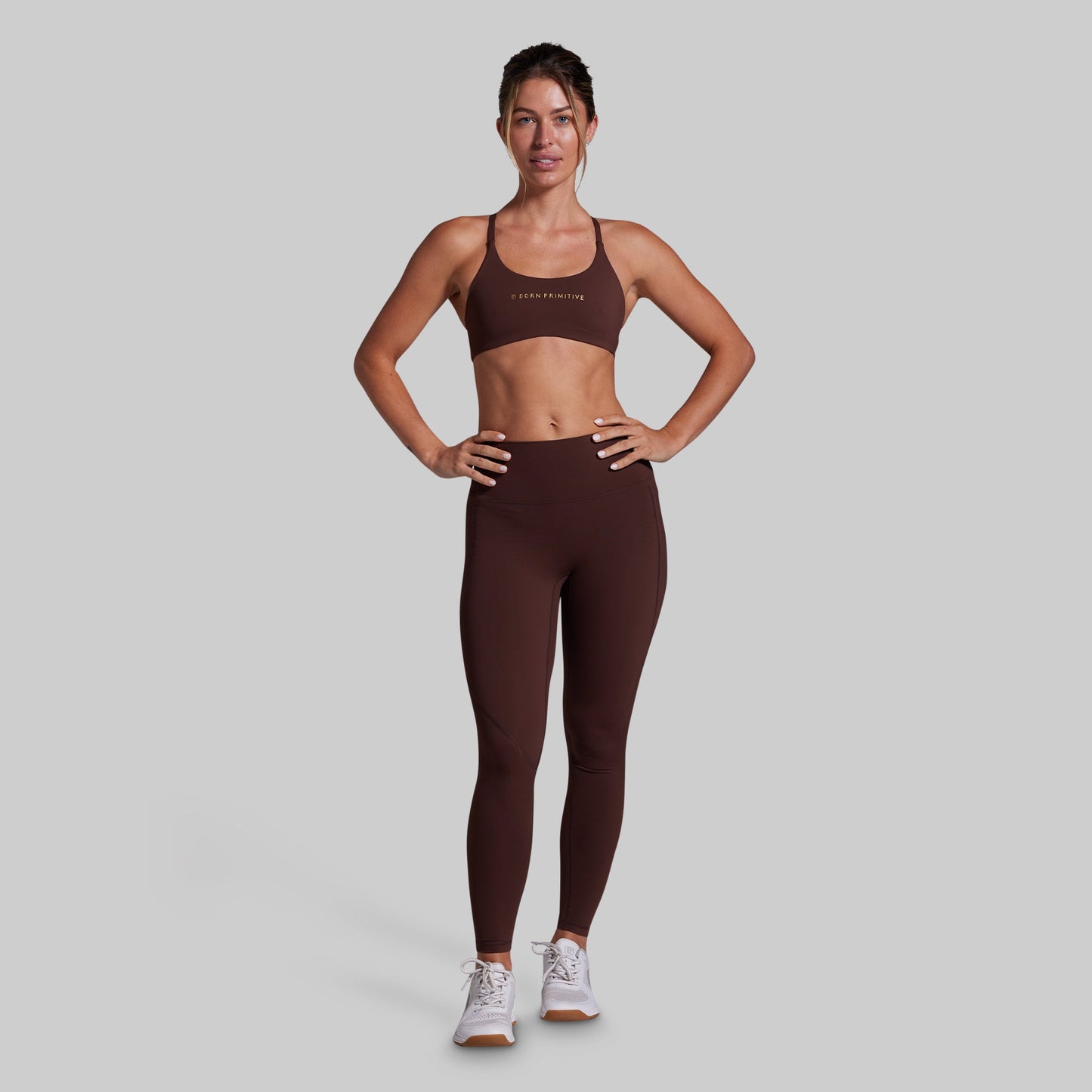 A woman modeling a matching dark brown activewear set consisting of a sports bra and high-waisted leggings, standing against a solid light gray background.