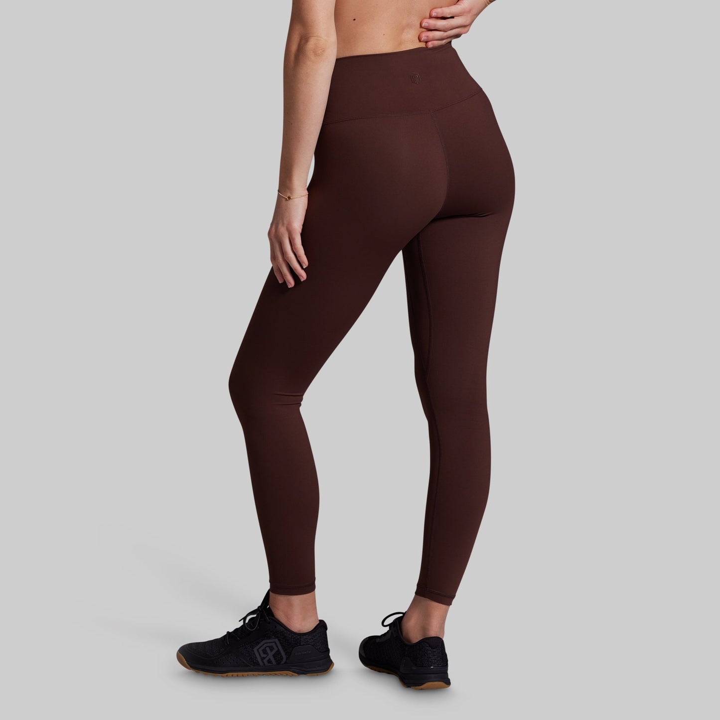 A model from the waist down wearing high-waisted, form-fitting leggings in a deep chocolate brown color, paired with black athletic shoes, against a solid light gray background.