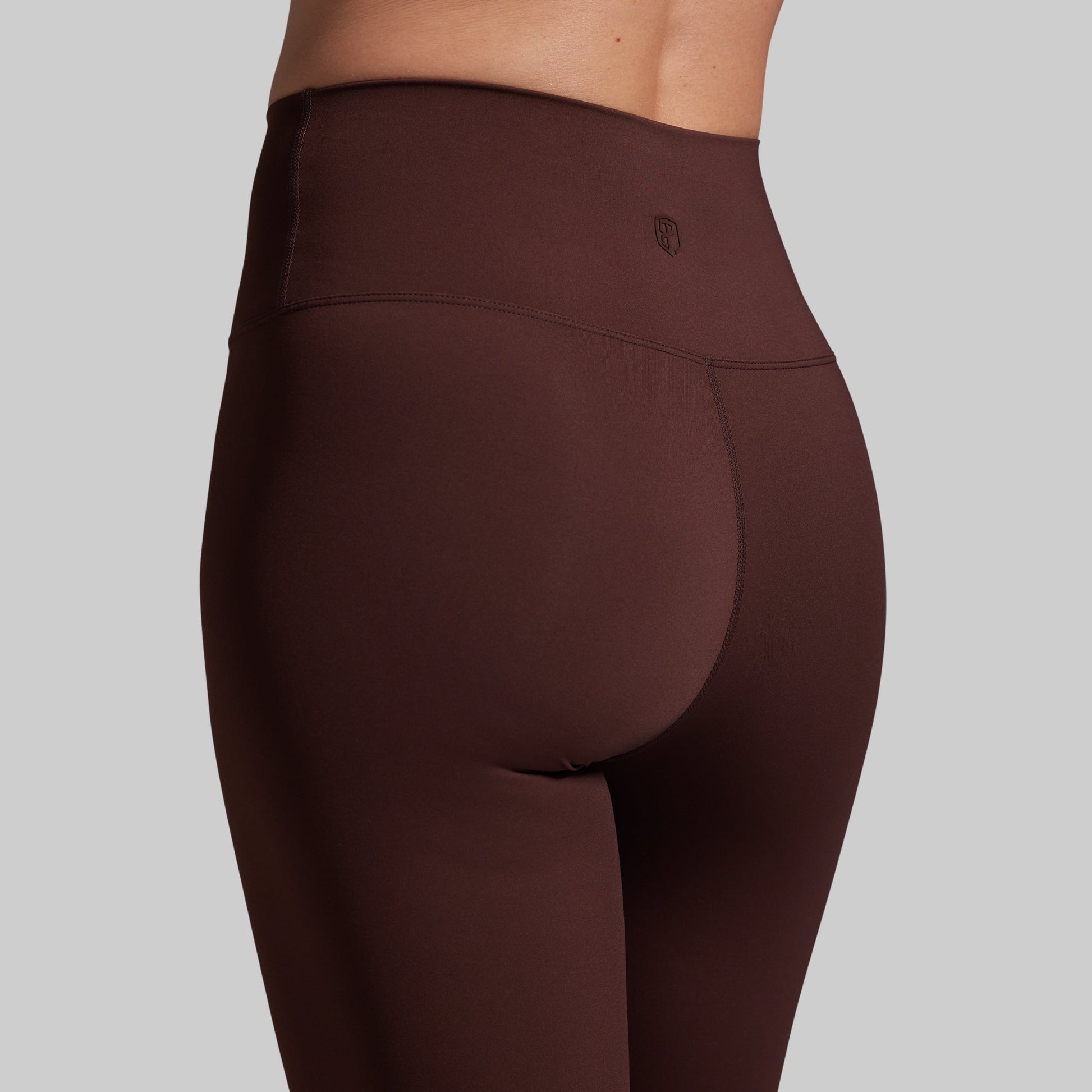 Rear view of a person wearing high-waisted leggings in a dark brown color, shown against a solid gray background.