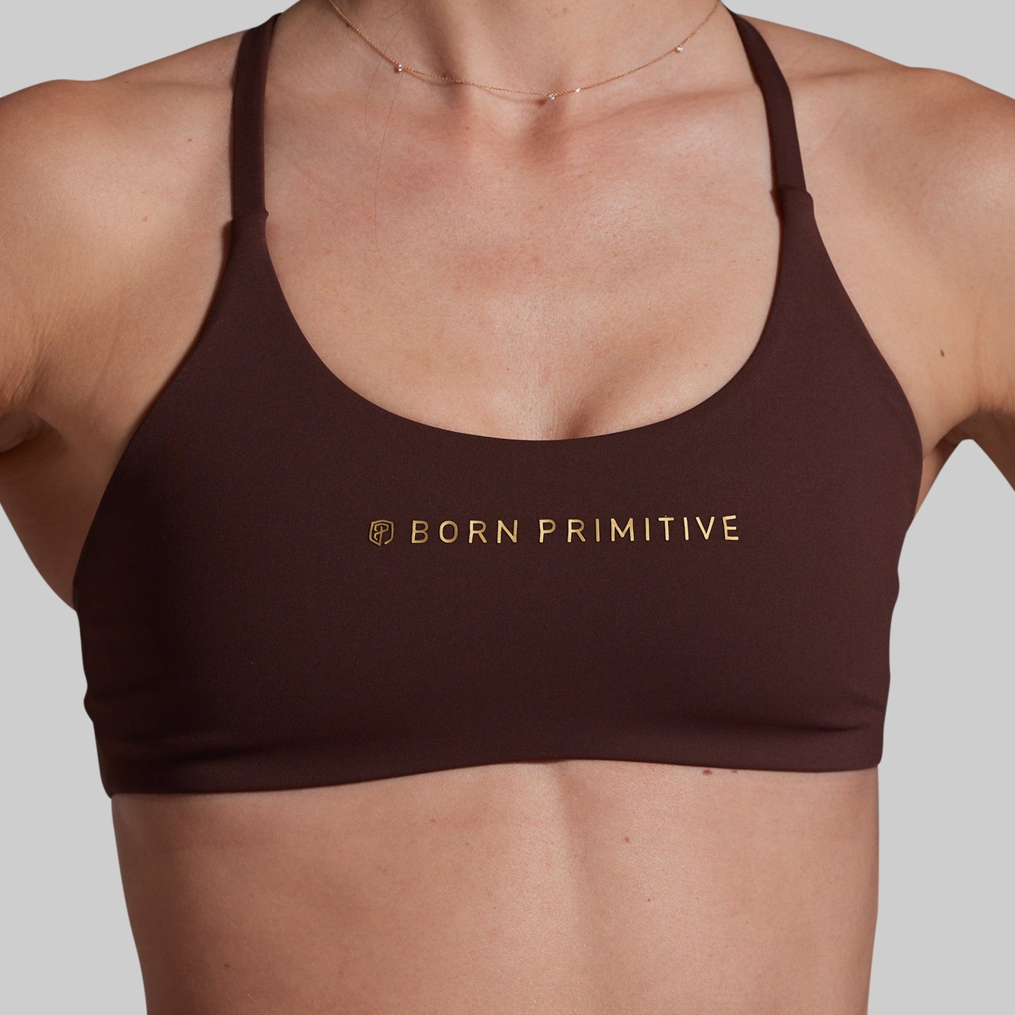 A close-up of a model wearing a dark brown sports bra with gold 'BORN PRIMITIVE' branding on the chest, set against a solid light gray background.