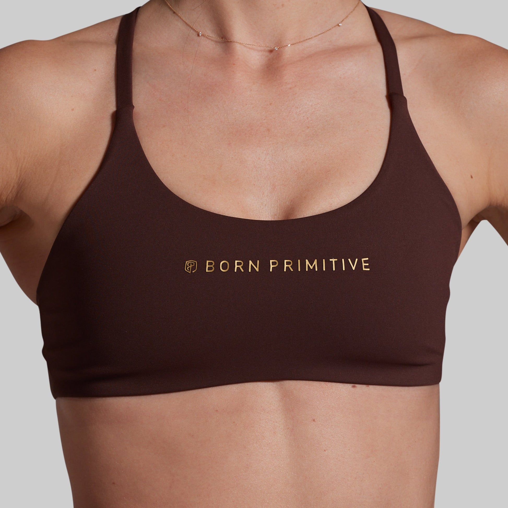 A close-up of a model wearing a dark brown sports bra with gold 'BORN PRIMITIVE' branding on the chest, set against a solid light gray background.