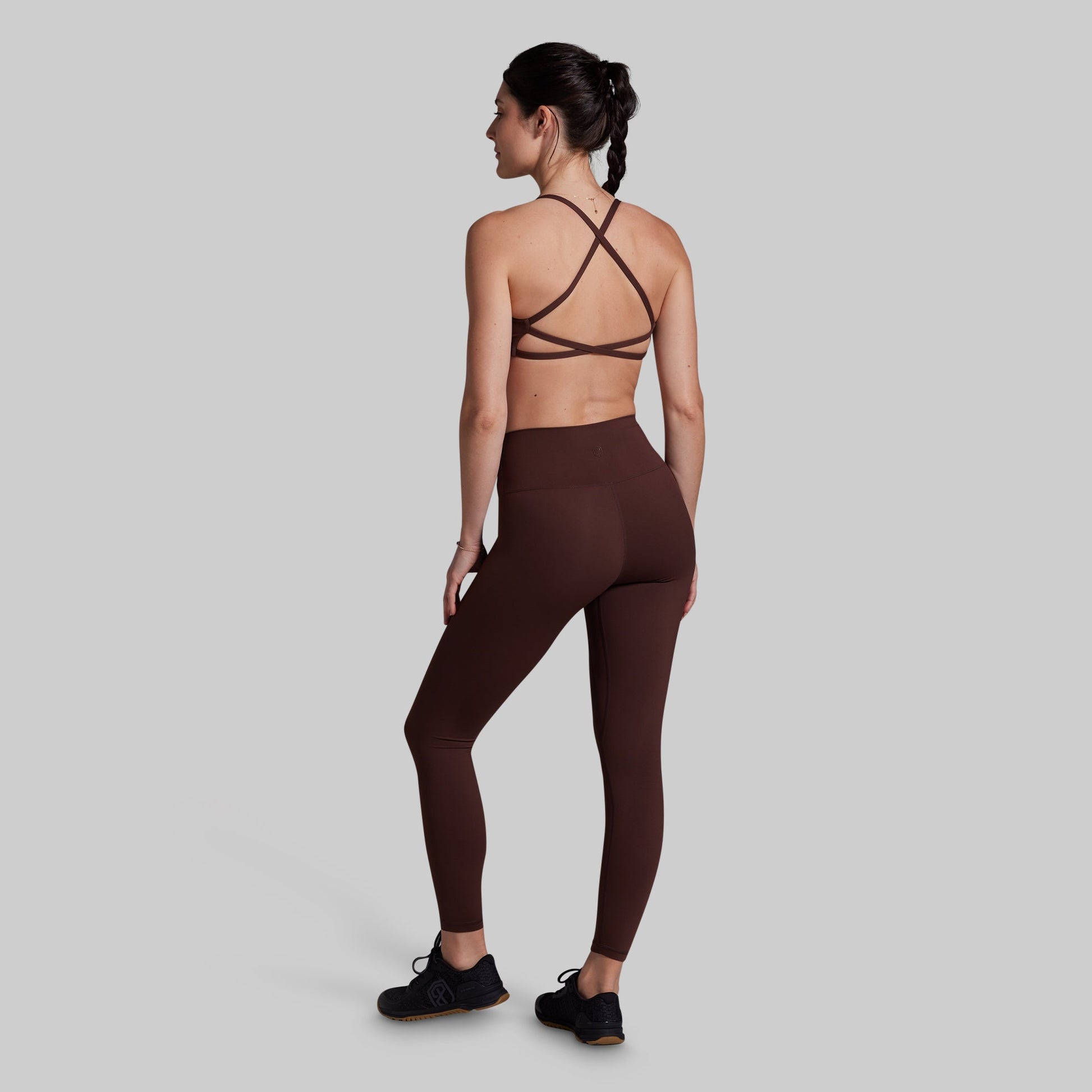 A woman shown from the back wearing a dark brown strappy sports bra and matching high-waisted leggings, paired with black sneakers, against a solid light gray background.