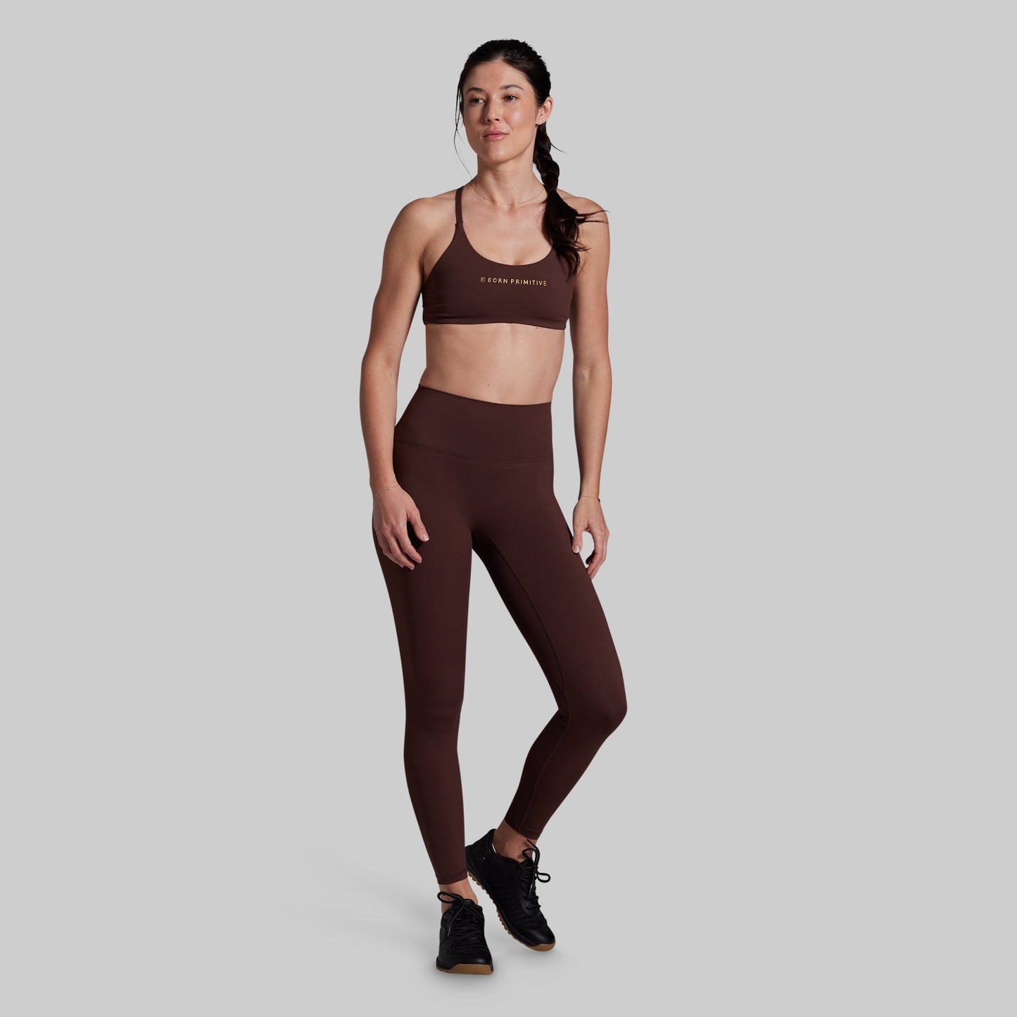 A woman modeling a matching dark brown activewear set consisting of a sports bra and high-waisted leggings, standing against a solid light gray background.