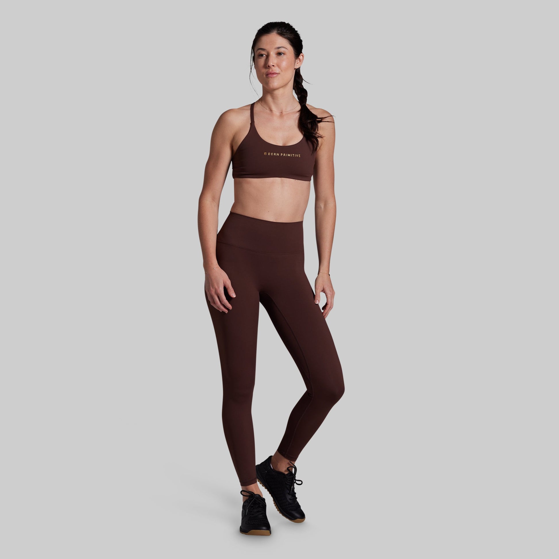 A woman modeling a matching dark brown activewear set consisting of a sports bra and high-waisted leggings, standing against a solid light gray background.