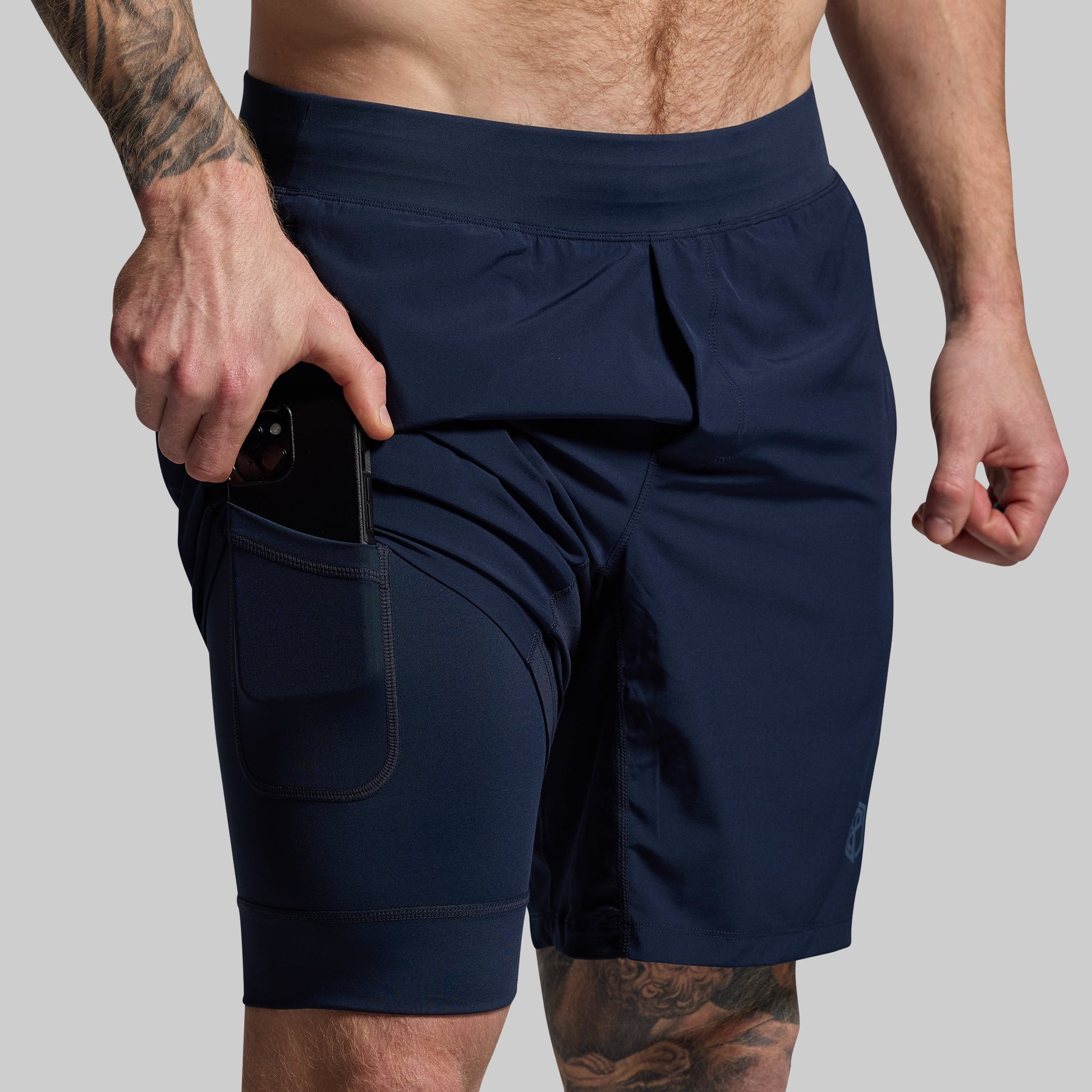 A man wearing navy blue athletic shorts with pockets and a visible mobile phone in one of the pockets on a compression liner.