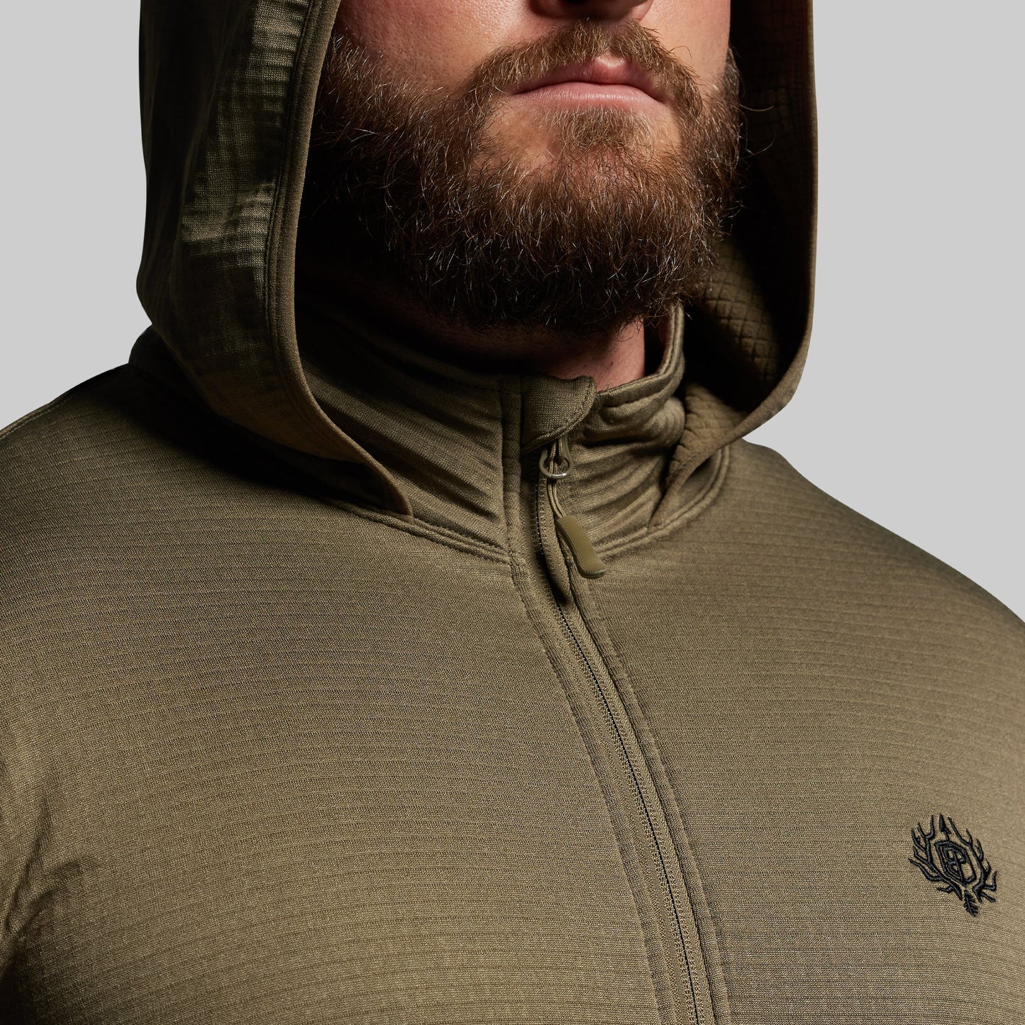 Person wearing a green hooded mid layer with a brand logo on a gray background