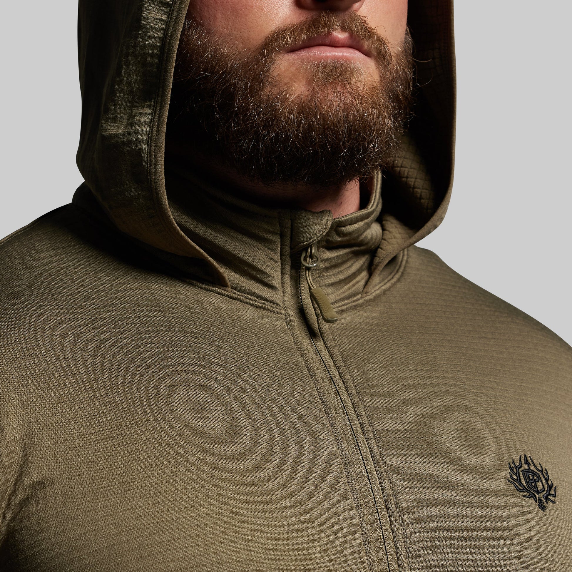 Person wearing a green hooded mid layer with a brand logo on a gray background