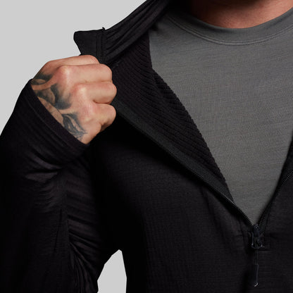 Person wearing a black quarter zip hoodie over a gray shirt with a tattooed arm.