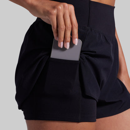 Person wearing black shorts with a pocket holding a phone on a plain background
