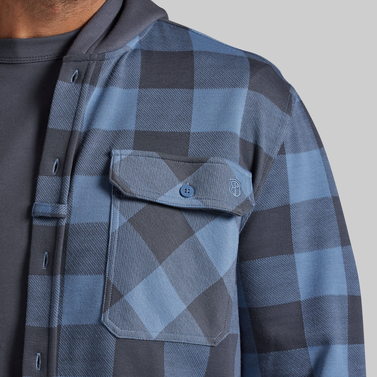 Close-up of a person wearing a blue and gray plaid overshirt over a dark gray shirt, showing the chest pocket and button closure against a plain light gray background.
