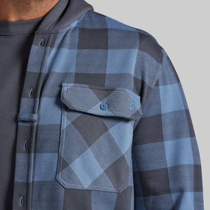 Close-up of a person wearing a blue and gray plaid overshirt over a dark gray shirt, showing the chest pocket and button closure against a plain light gray background.