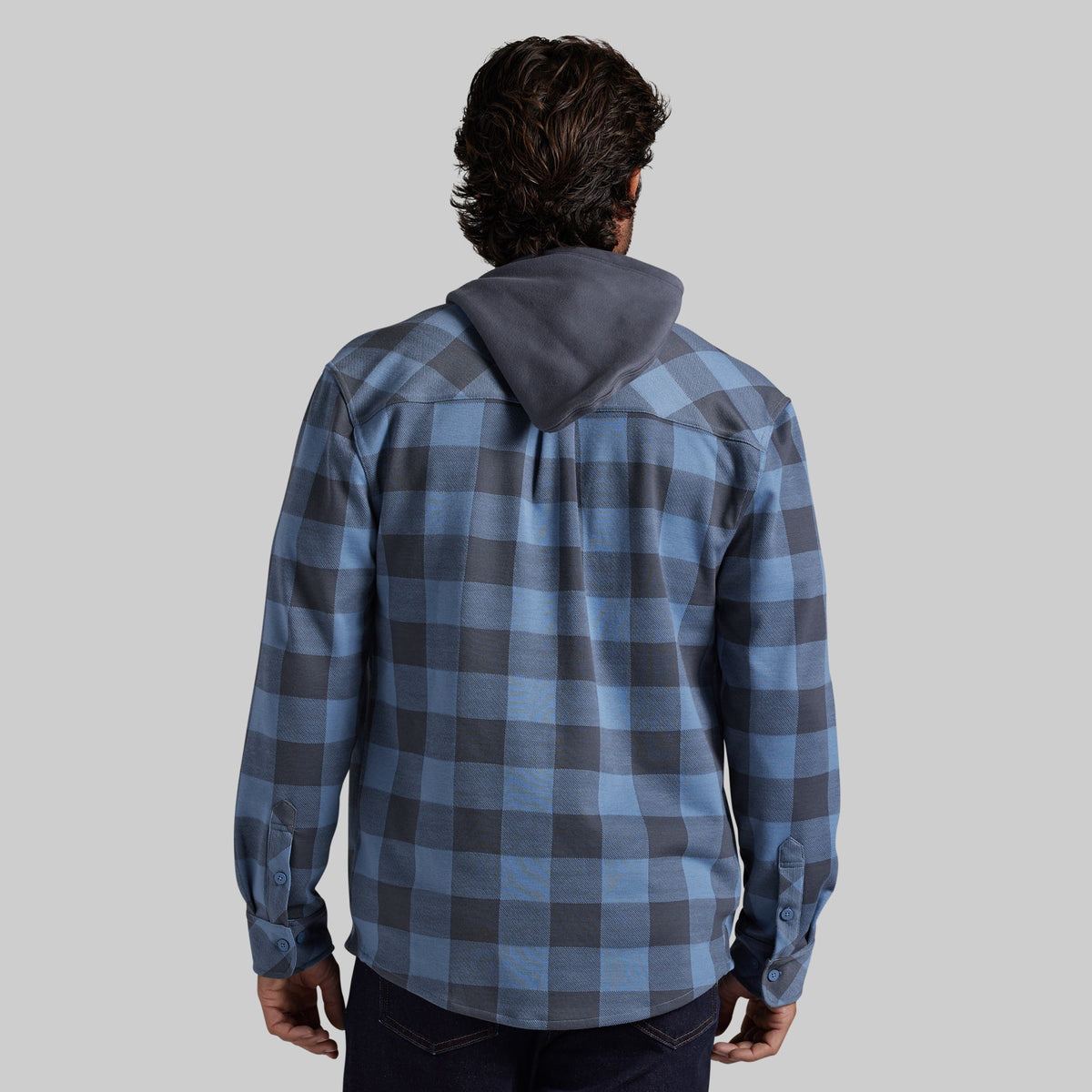 A man viewed from behind wearing a blue and gray plaid hooded shirt, set against a solid light gray background.