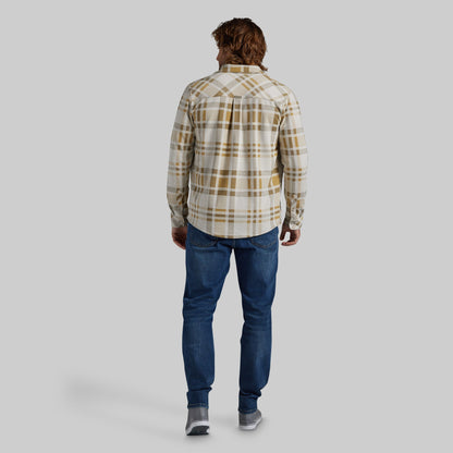 A man viewed from behind wearing a cream and tan plaid button-down shirt, blue jeans, and gray sneakers, standing against a solid light gray background.