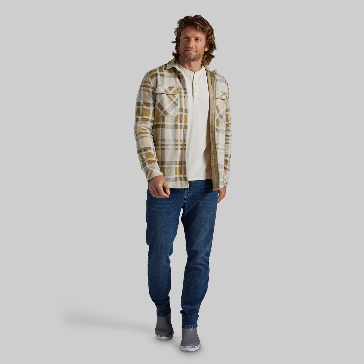 A man modeling a long-sleeve plaid flannel shirt in beige and brown over a white henley t-shirt, paired with blue jeans and gray boots, against a solid gray background.