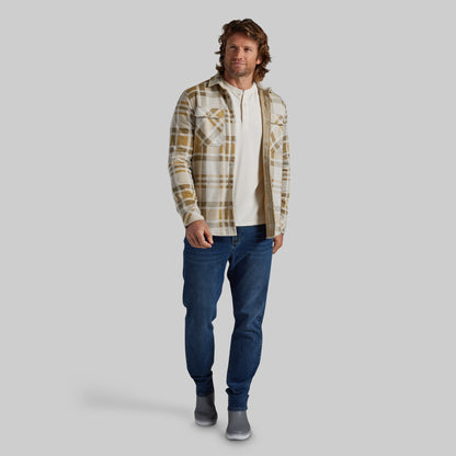 A man modeling a long-sleeve plaid flannel shirt in beige and brown over a white henley t-shirt, paired with blue jeans and gray boots, against a solid gray background.