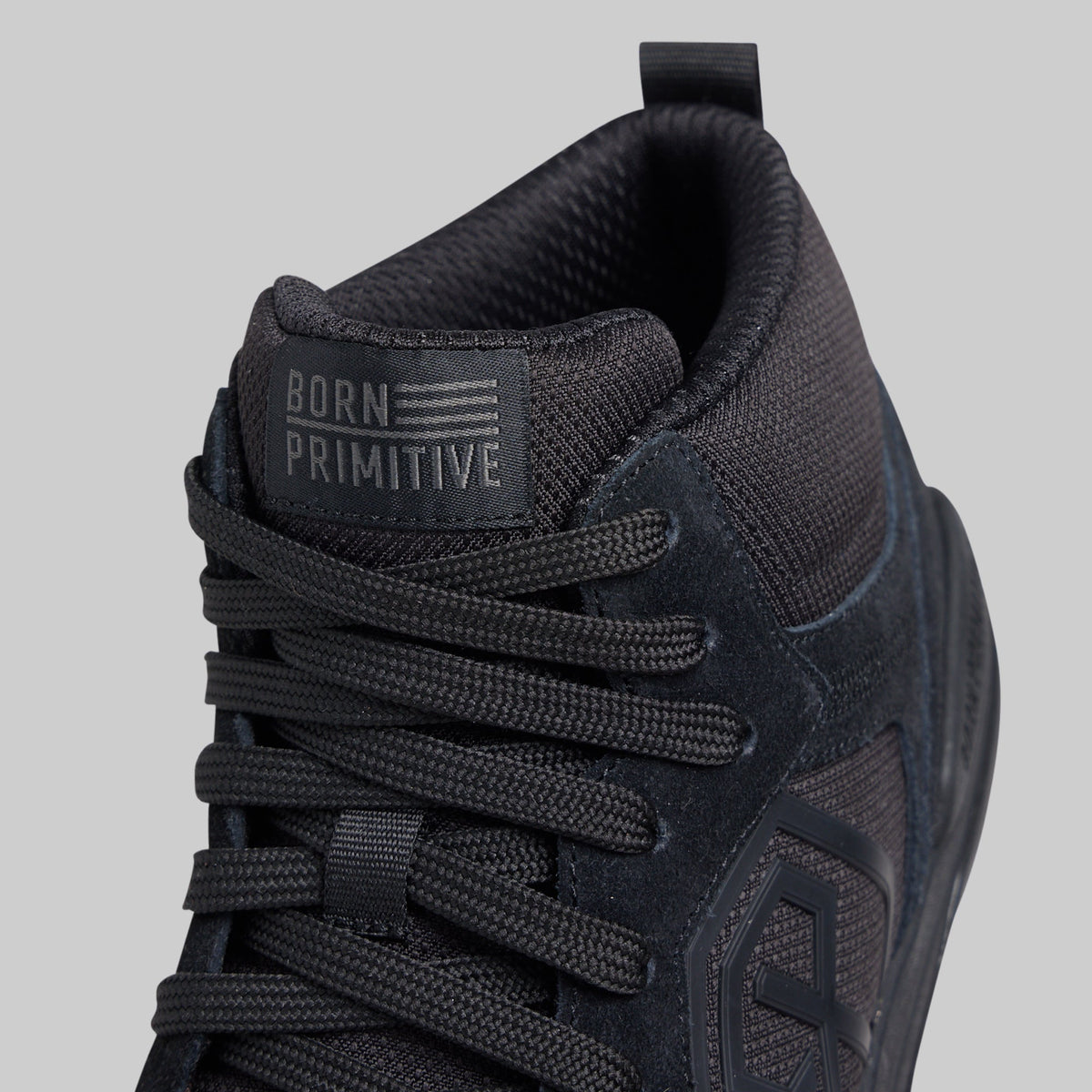 Close-up of a black mid-top sneaker by Born Primitive, showing the textured mesh and suede upper, black laces, and the brand's tongue label.