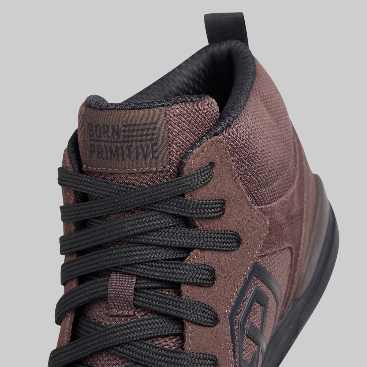 Close-up of a mid-top sneaker by Born Primitive, featuring a textured brown and black upper with black laces and a black pull tab, set against a solid gray background.