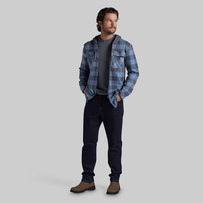 A man modeling a blue and gray plaid hooded flannel shirt over a dark gray t-shirt, dark jeans, and brown boots, standing against a solid gray studio background.