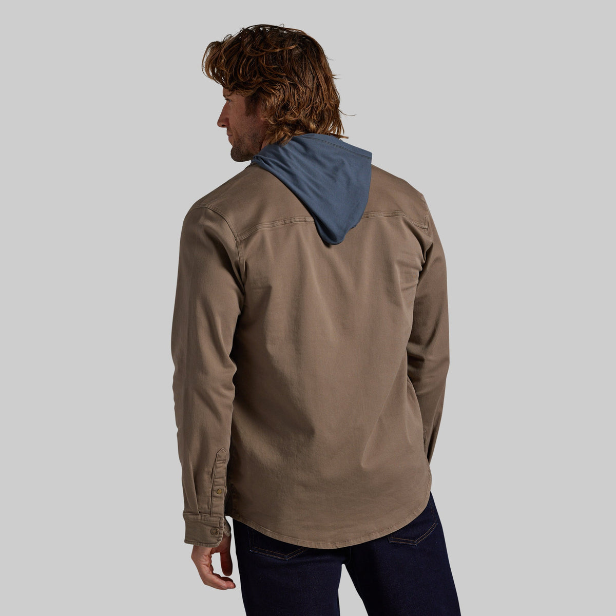 A man shown from the back wearing a brown shirt jacket with a contrasting blue hood, paired with dark blue jeans, against a solid gray background.