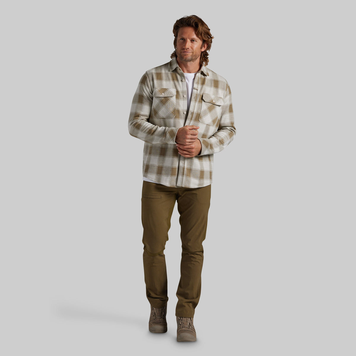 A man wearing a beige and brown plaid flannel shirt over a white undershirt, paired with olive green pants, set against a solid gray background.