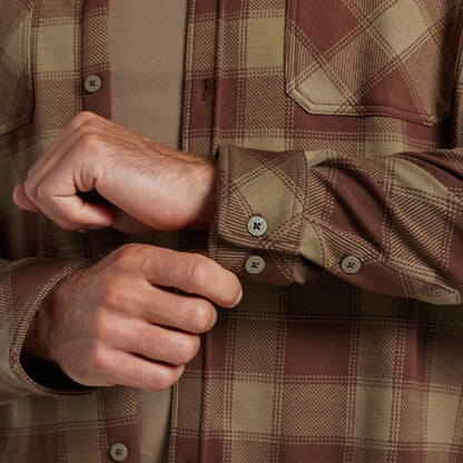 Close-up of a man's hands buttoning the cuffs of a brown and tan plaid flannel shirt over a tan undershirt.