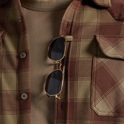 Close-up of a person wearing a brown and tan plaid flannel shirt over a taupe t-shirt, with gold-framed sunglasses hanging from the placket.