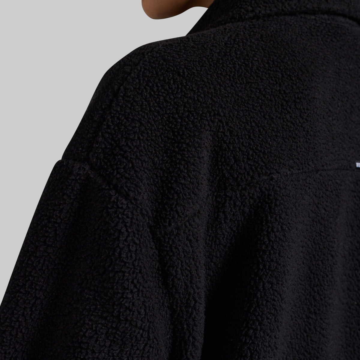 Close-up of a person wearing a black textured fleece jacket, showing the collar, shoulder, and fabric texture against a light gray background.