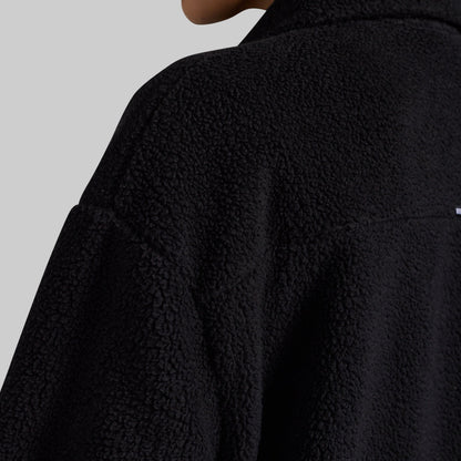 Close-up of a person wearing a black textured fleece jacket, showing the collar, shoulder, and fabric texture against a light gray background.
